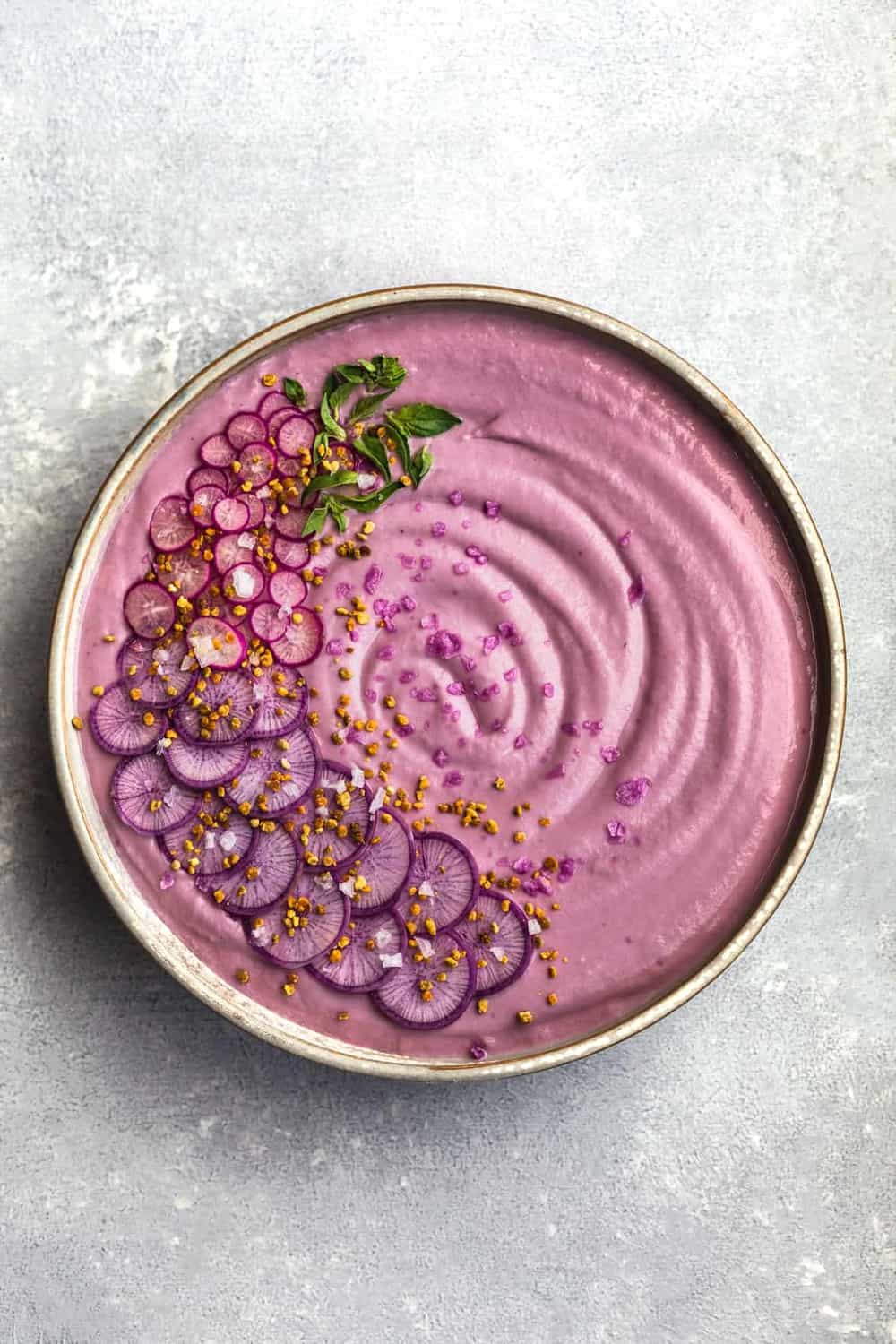 Vegan Purple Cauliflower Soup with Sliced Purple and Pink Radishes, tiny basil leaves, Bee Pollen and Flaky Sea Salt. Overhead shot. Bowl is in center of the frame on a white/light blue background.