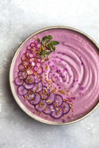 VeganPurple Cauliflower Soup with Sliced Purple and Pink Radishes, tiny basil leaves, Bee Pollen and Flaky Sea Salt. Overhead shot, right side cut off, on a white/light blue background.