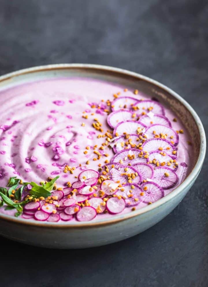 Vegan Purple Cauliflower Soup with Sliced Purple and Pink Radishes, tiny basil leaves, Bee Pollen and Flaky Sea Salt. Side angle shot, left side cut off, on a dark grey white/light blue background.