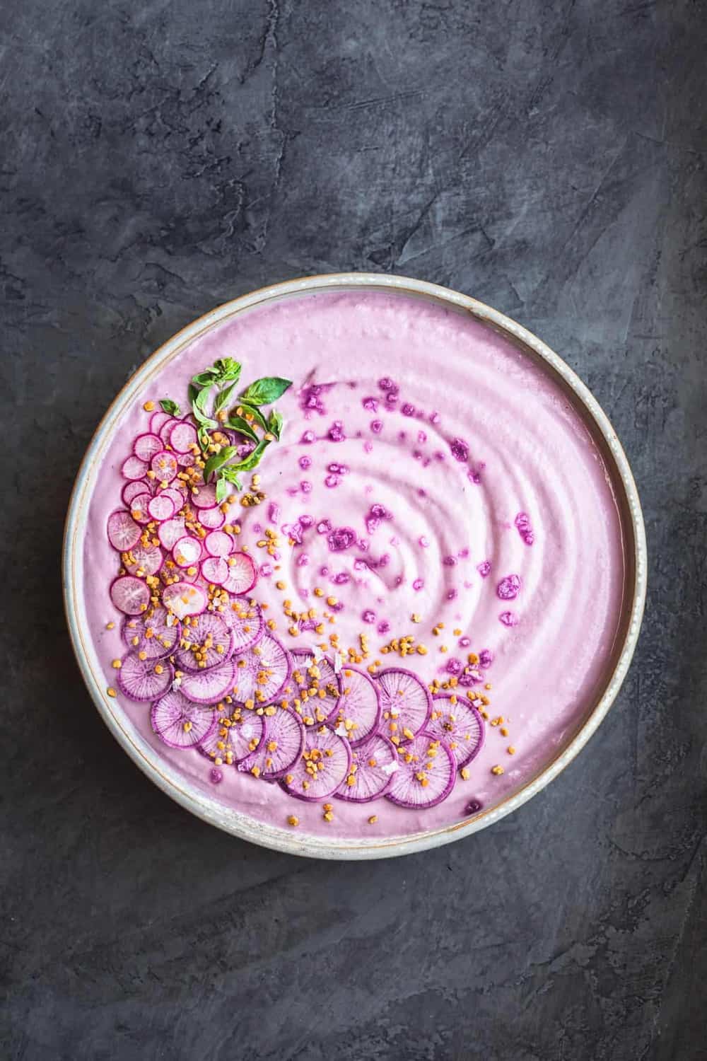 Vegan Purple Cauliflower Soup with Sliced Purple and Pink Radishes, tiny basil leaves, Bee Pollen and Flaky Sea Salt. Overhead shot. Bowl is in center of the frame on a dark grey background.