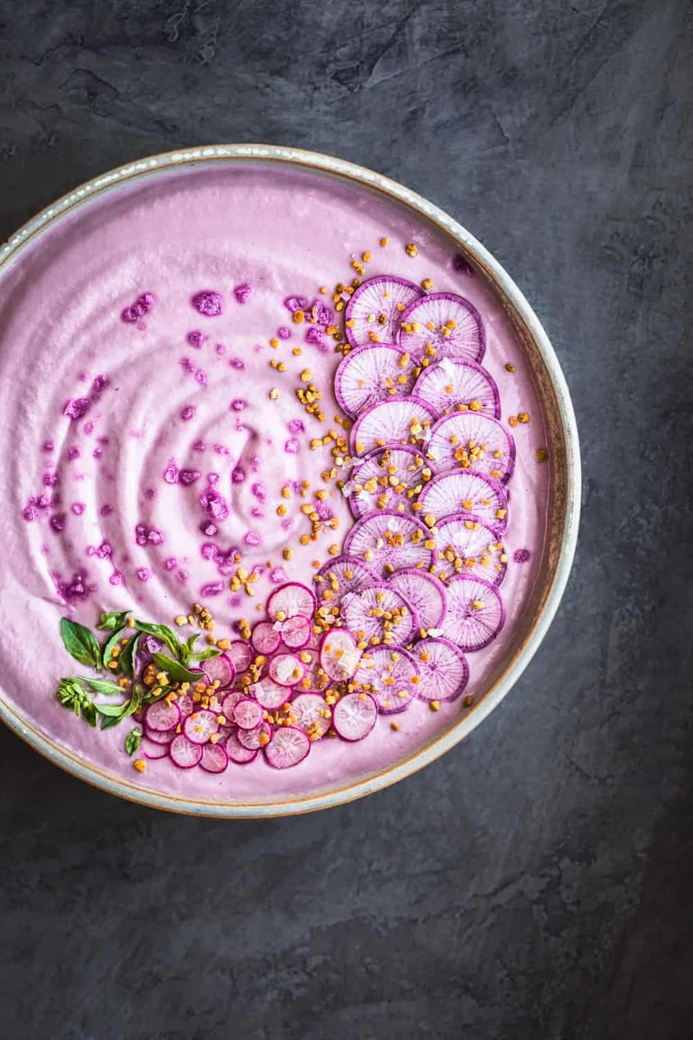 Vegan Purple Cauliflower Soup with Sliced Purple and Pink Radishes, tiny basil leaves, Bee Pollen and Flaky Sea Salt. Overhead shot, left side of bowl is cut off, on a dark grey background.