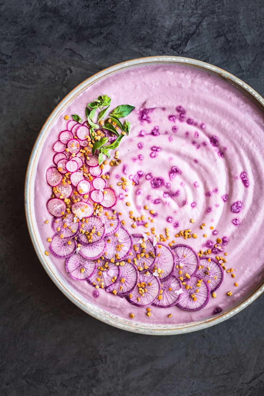 Vegan Purple Cauliflower Soup with Sliced Purple and Pink Radishes, tiny basil leaves, Bee Pollen and Flaky Sea Salt. Overhead shot, right side of bowl is cut off, on a dark grey background.