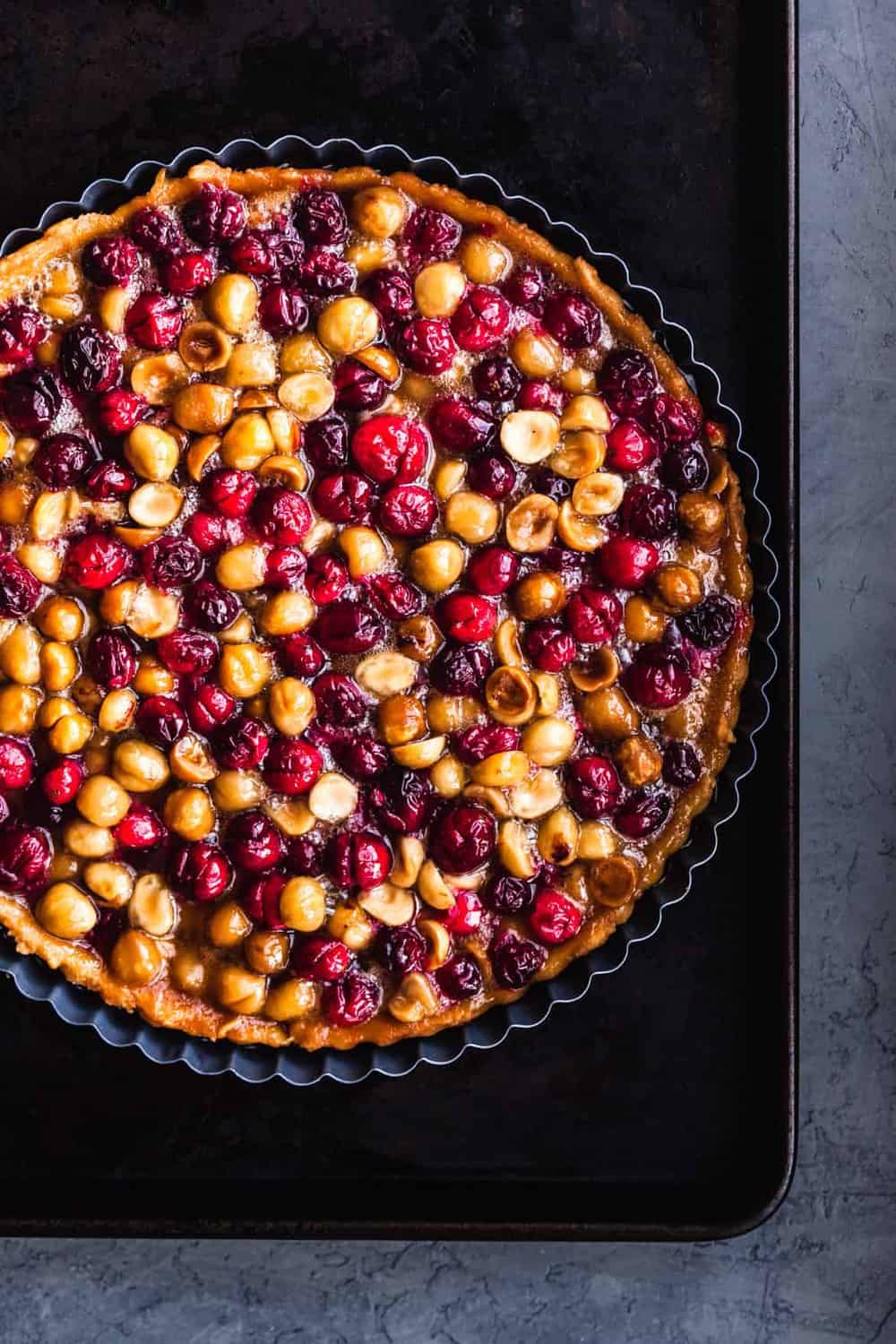 Candied Macadamia, Cranberry & Caramel Tart on a sheet pan, over head, on black surface, with the right side cut off.