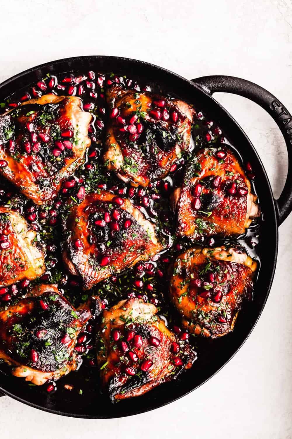 Pomegranate Chicken topped with pomegranate arils and parsley, in cast iron skillet on a white background; overhead shot.