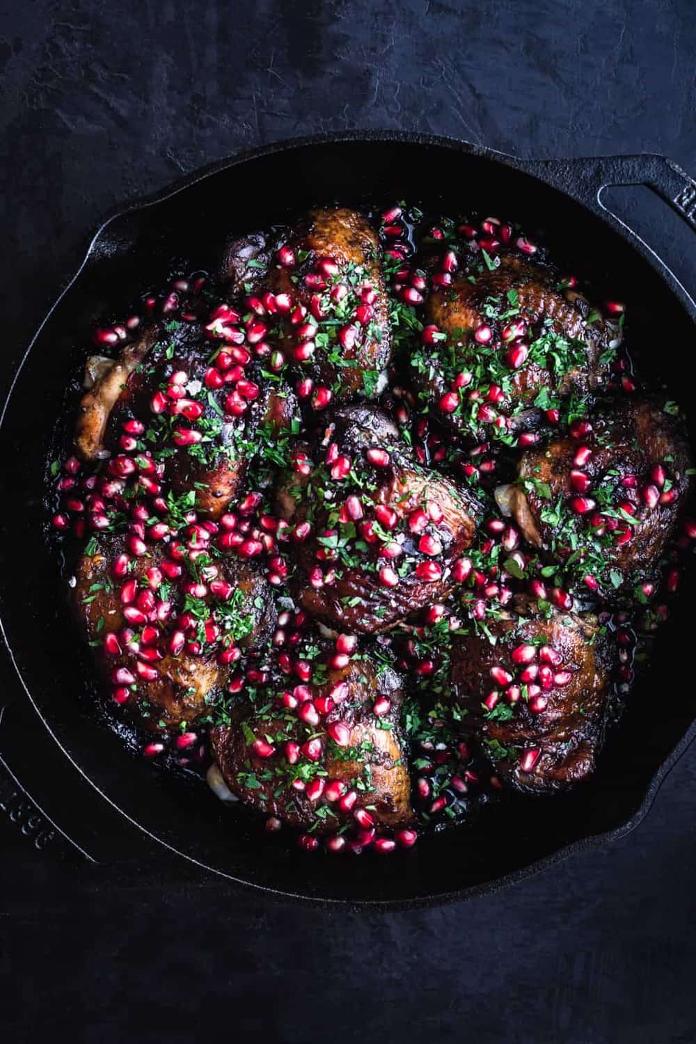 Pomegranate Chicken, all done sprinkled with parsley and fresh pomegranate arils. In a cast iron skillet on a black background; overhead shot.