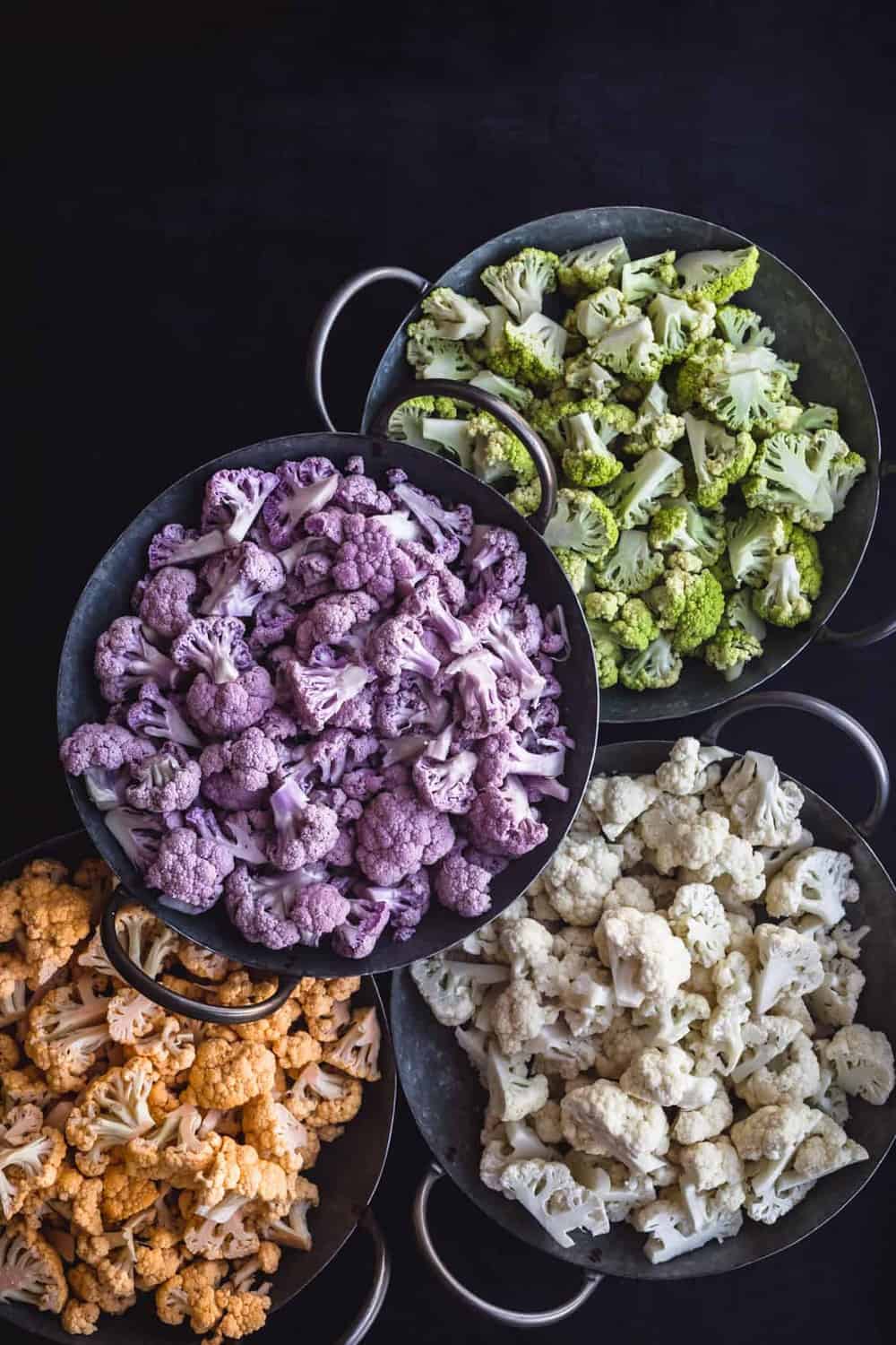 Yellow/Orange, Purple, White, and Green Cauliflower, each cut into florets and separated by color into 4 silvers bowls with handles, on a black background.