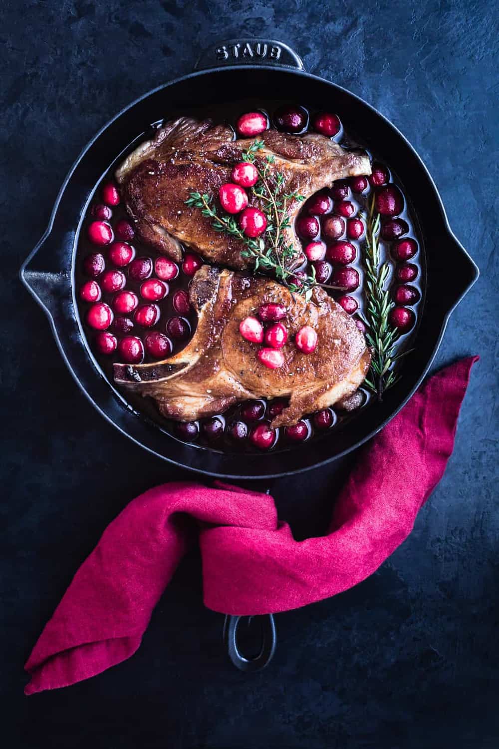 Pork chops have been seared and are in a cast iron skillet with the cranberry sauce all ready for the oven. Overhead shot with a red linen around the pan handle on a black background.