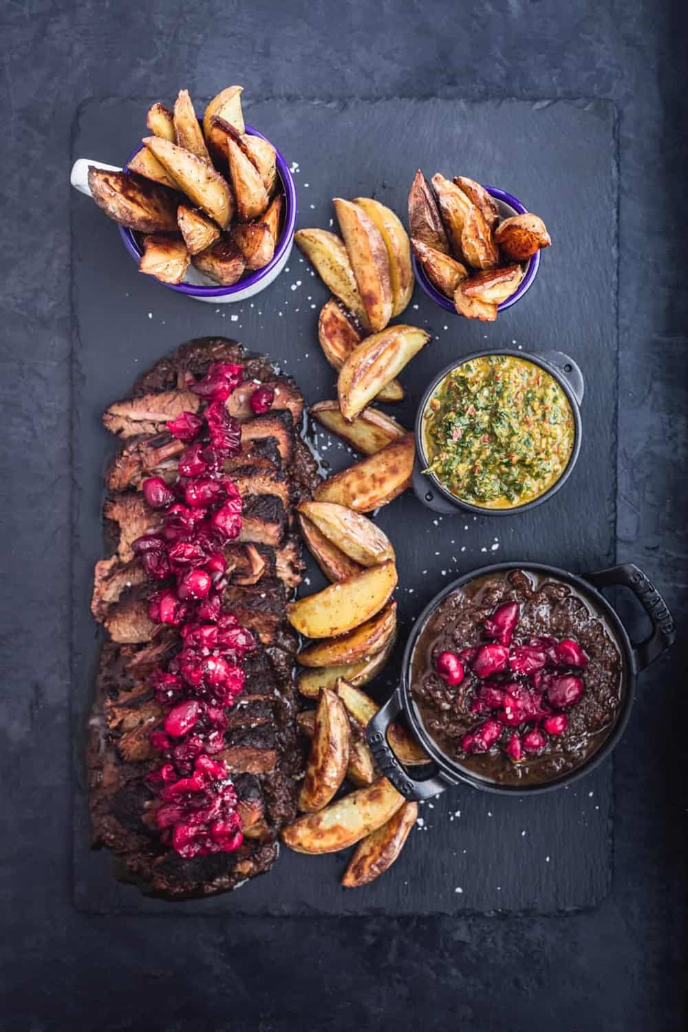 Thinly sliced brisket on a black slate with cranberry sauce on top, baked wedge potatoes and 2 little bowls of chimichurri and more cranberry sauce. Overhead shot on a back background.
