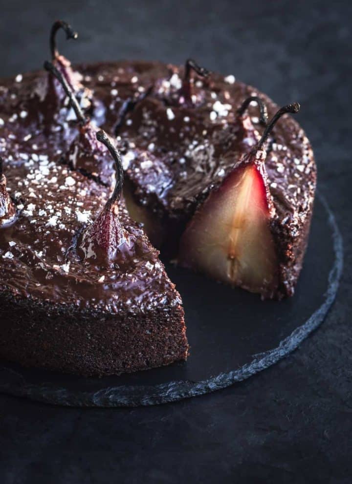 Spiced red wine poached pears in chocolate cake batter, out of cake pan, post oven, topped with ganache and flaky sea salt. Side angle shot. Cake is cut into and can see a full pear sliced, on black background, and left side of cake is cut off.
