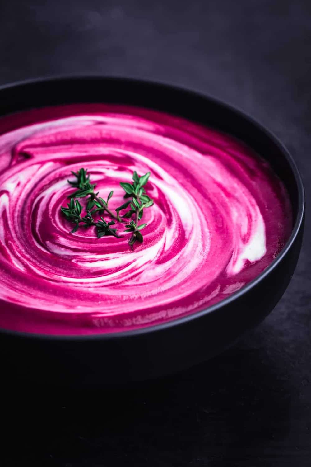 Roasted Beet Soup with Cashew Cream swirl and thyme sprigs, side angle shot on a black background.