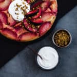 A stylized food shot of the plum upside down cake, a bowl of yogurt and a bowl of pistachios.