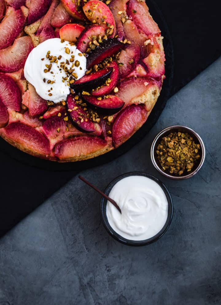 A stylized food shot of the plum upside down cake, a bowl of yogurt and a bowl of pistachios.
