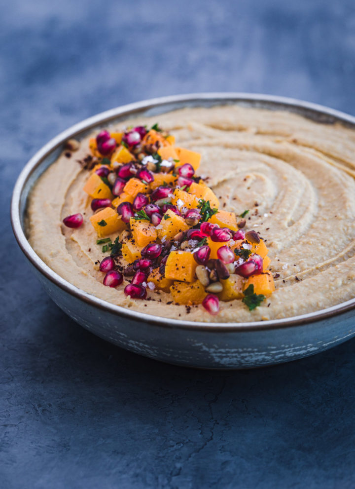 Side angle shot of butternut squash roasted hummus with cubed butternut roasted squash, pomegranate seeds, pastel, pinuts, sumac and flaky sea salt on top.