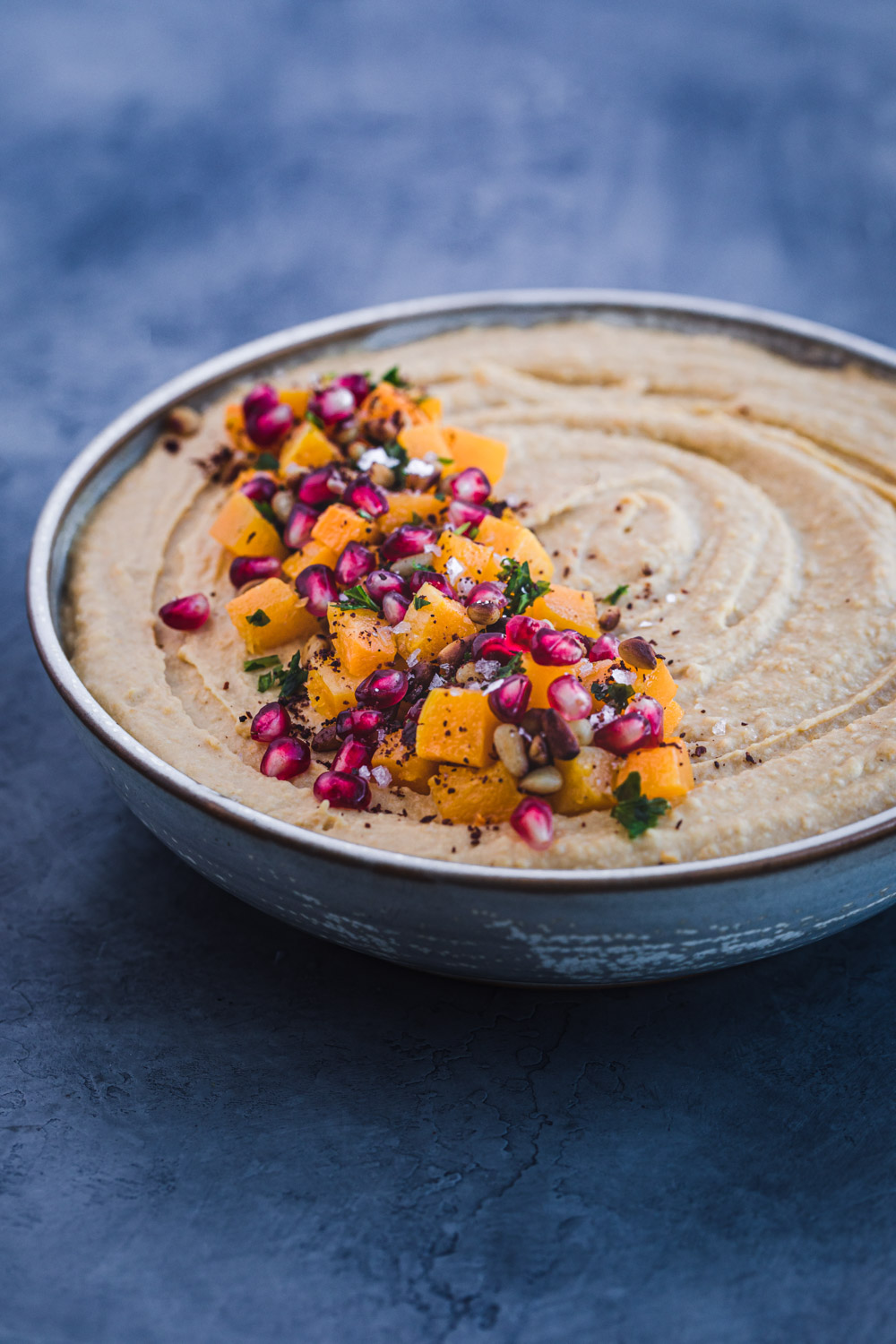Side angle shot of butternut squash roasted hummus with cubed butternut roasted squash, pomegranate seeds, pastel, pinuts, sumac and flaky sea salt on top.