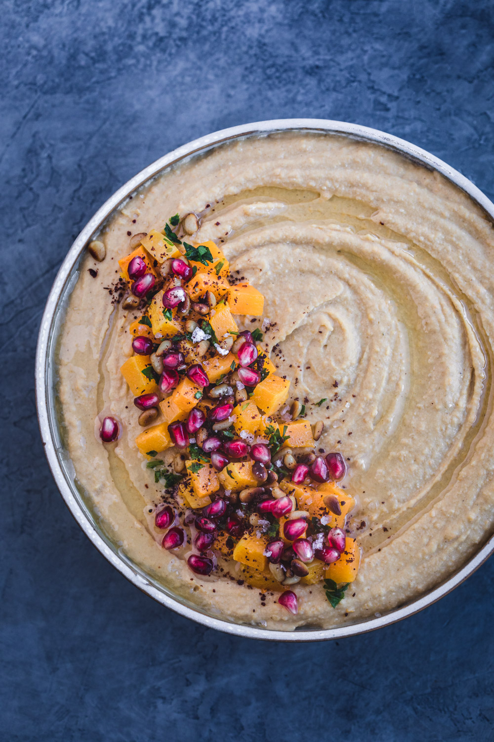 Overshot shot of butternut squash roasted hummus with cubed butternut roasted squash, pomegranate seeds, pastel, pinuts, sumac and flaky sea salt on top.