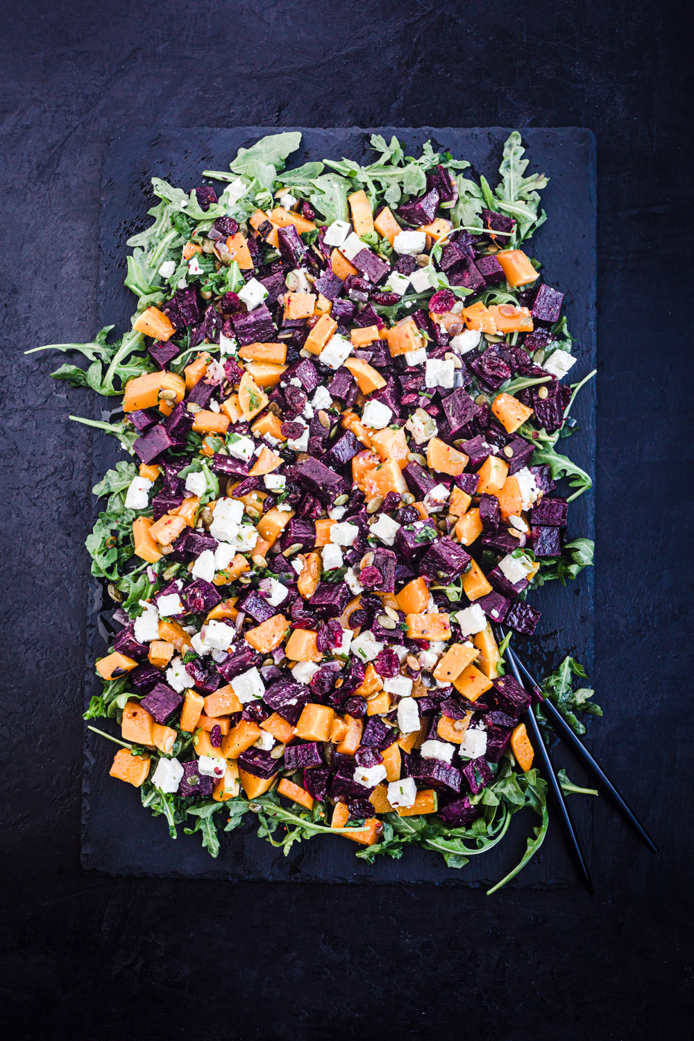 Finished sweet potato salad recipe shot, arranged on a black slate on a black background, with 2 black serving pieces in it.