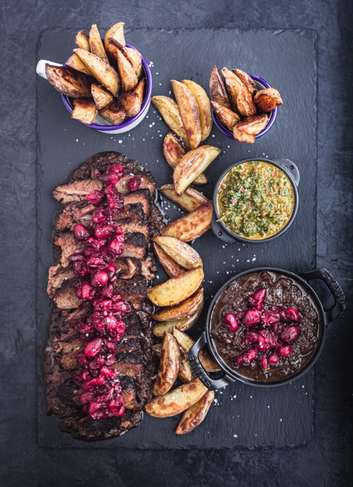 Thinly sliced brisket on a black slate with cranberry sauce on top, baked wedge potatoes and 2 little bowls of chimichurri and more cranberry sauce.