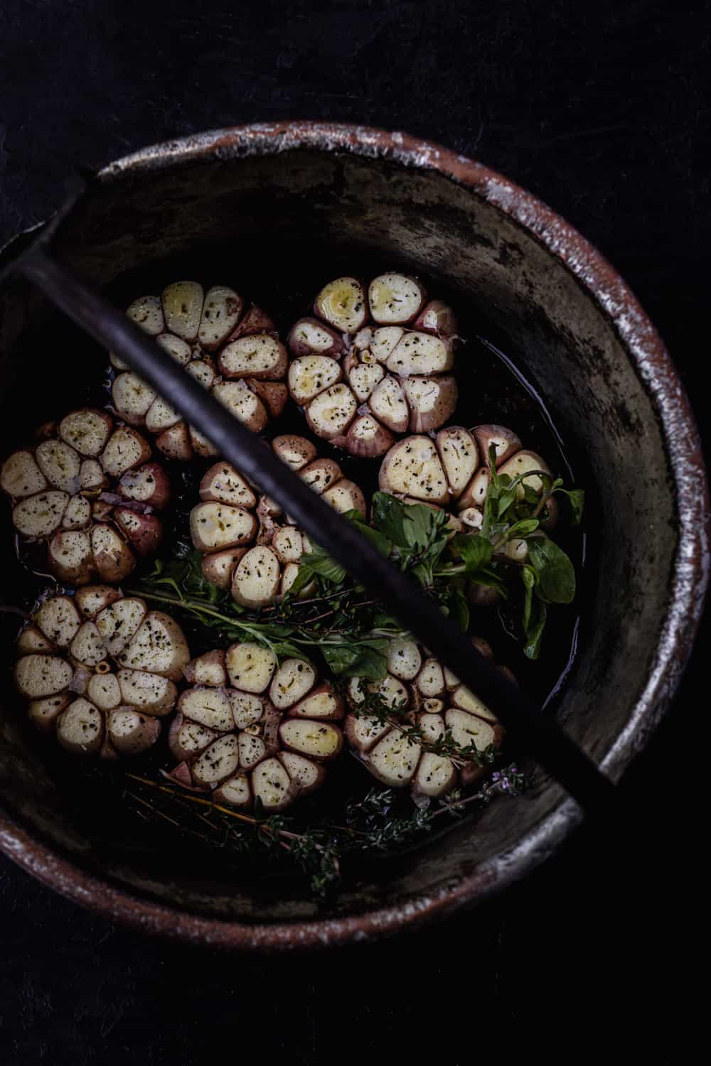 Overhead shot of a pot filled with full heads of garlic, tops trimmed off, with fresh herbs, salt and pepper.