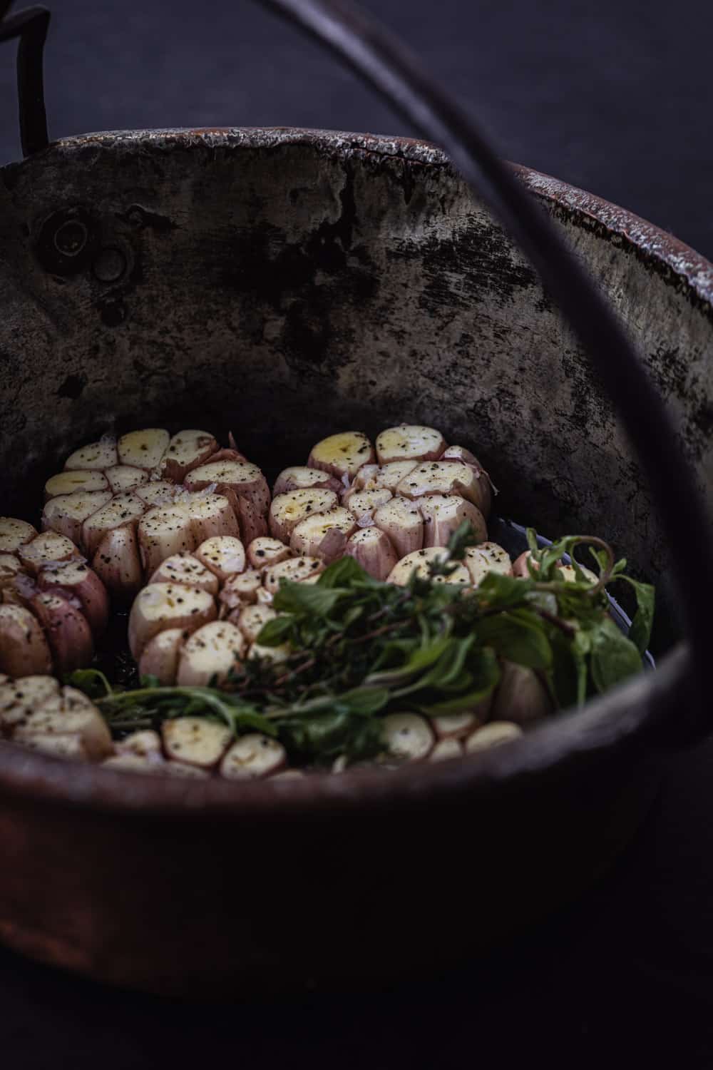 Prep shot of full heads of garlic, with the tops trimmed off and the cloves exposed, with fresh herbs in the pot too.