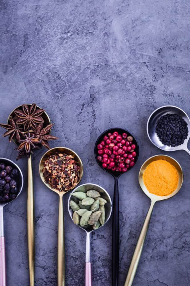 ingredient shot of 5 spices and black sesame seeds in spoons on a grey background