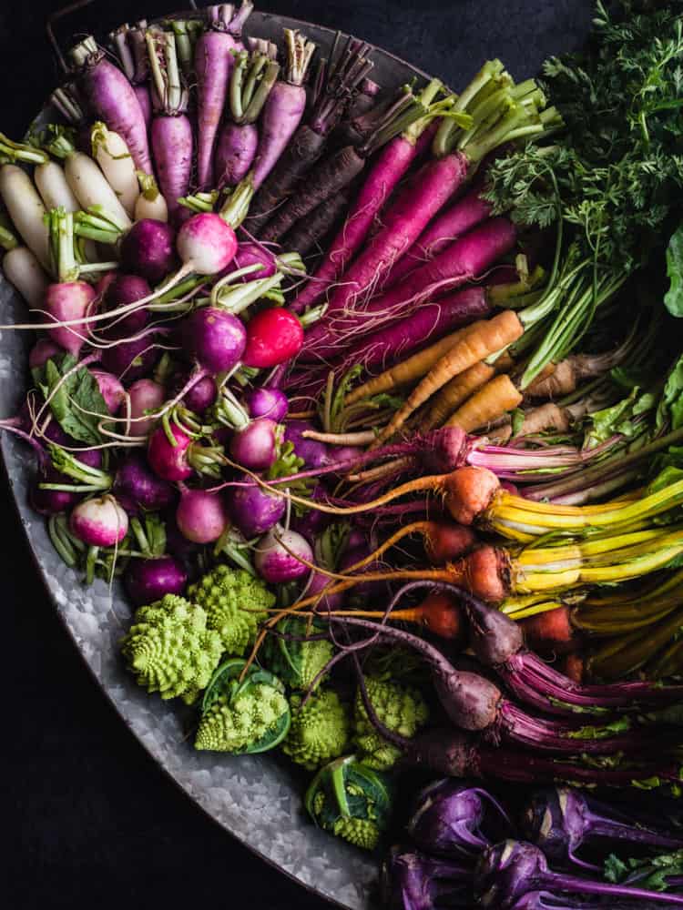 Platter filled with very colorful rbaby sized root veggies arranged like a rainbow; radishes, beets, kohlrabi, carrots and Romanesco.