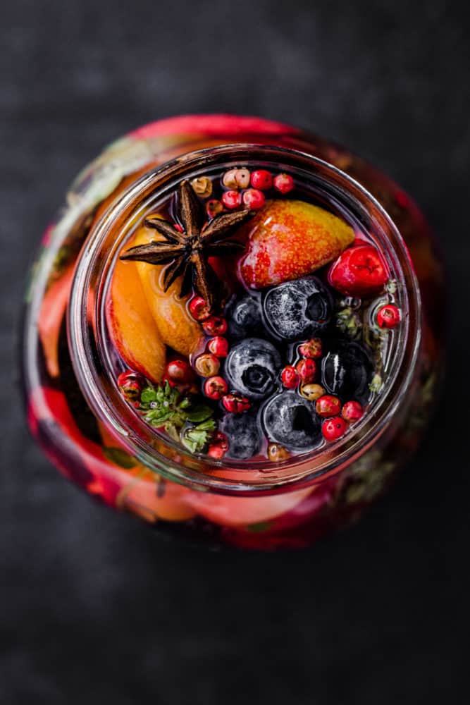 Blueberries, peaches and other fruit pickled in a jar with start anise, pink peppercorns, chili, thyme, and pickling brine, overhead shot.
