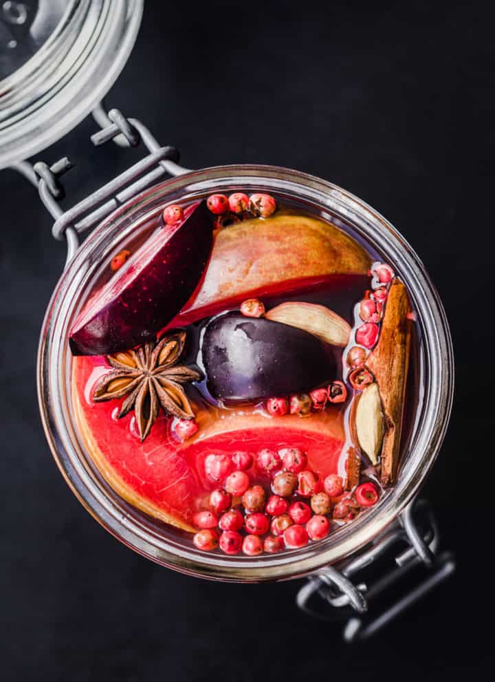 Pickled plums with star anise, cinnamon, pink peppercorns and pickling brine, overhead shot on a black background.