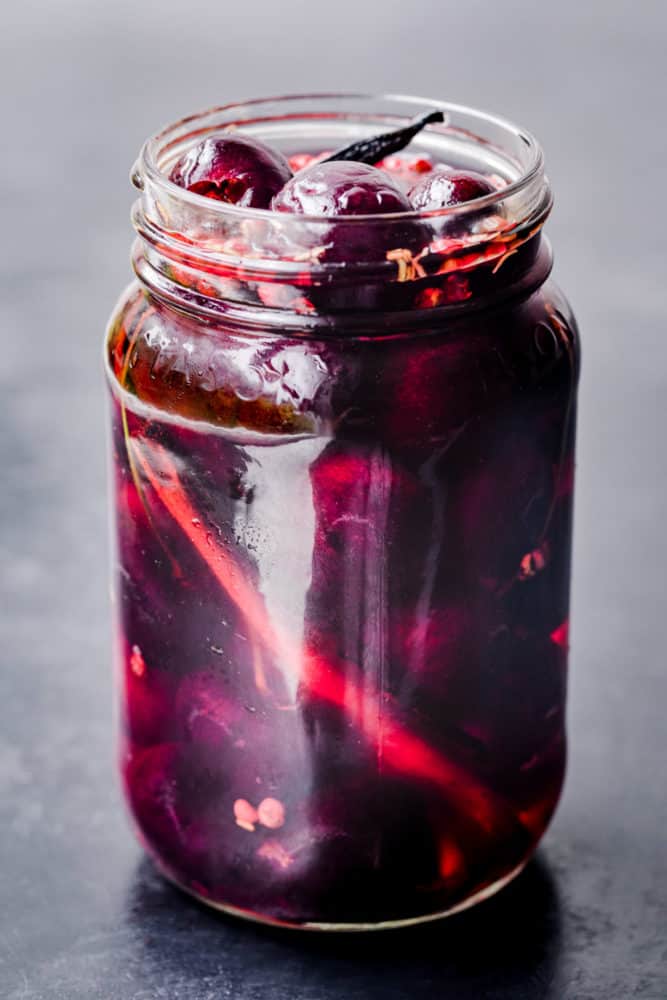 Pickled cherries! Cherries pickled in a jar with rosemary, vanilla, pink peppercorns, cinnamon and pickling brine, side angle shot.
