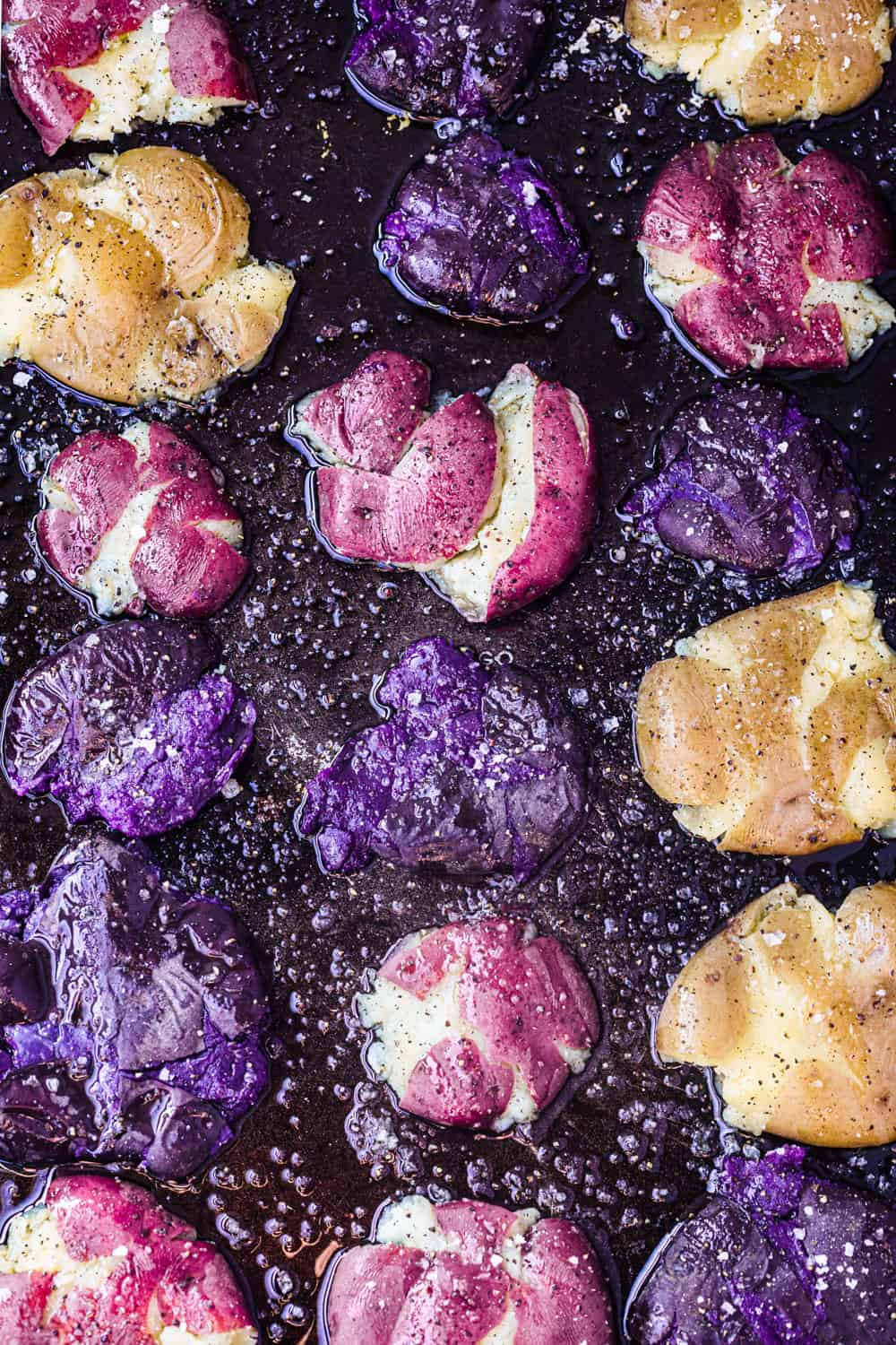 Smashed purple, pink and white potatoes drizzled with olive oil and flaky sea salt on black baking sheet; overhead shot.
