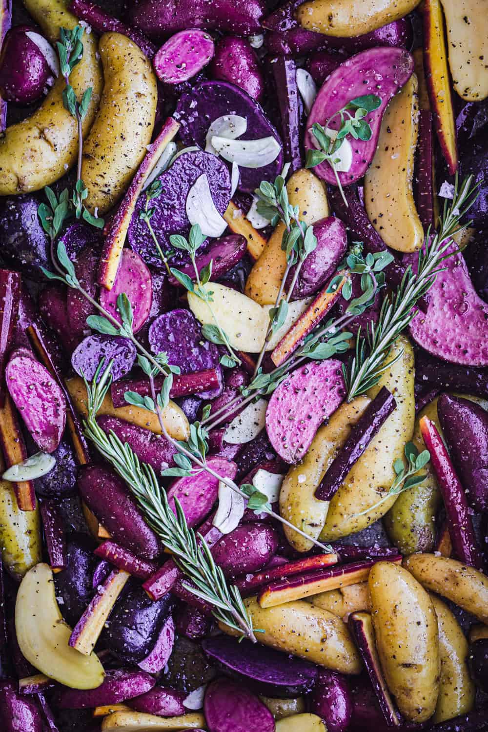 Purple, pink and white fingerling potatoes cut in half with olive oil, salt, pepper, rosemary, oregano, garlic and a few carrots, all mixed together on a sheet pan; overhead shot.