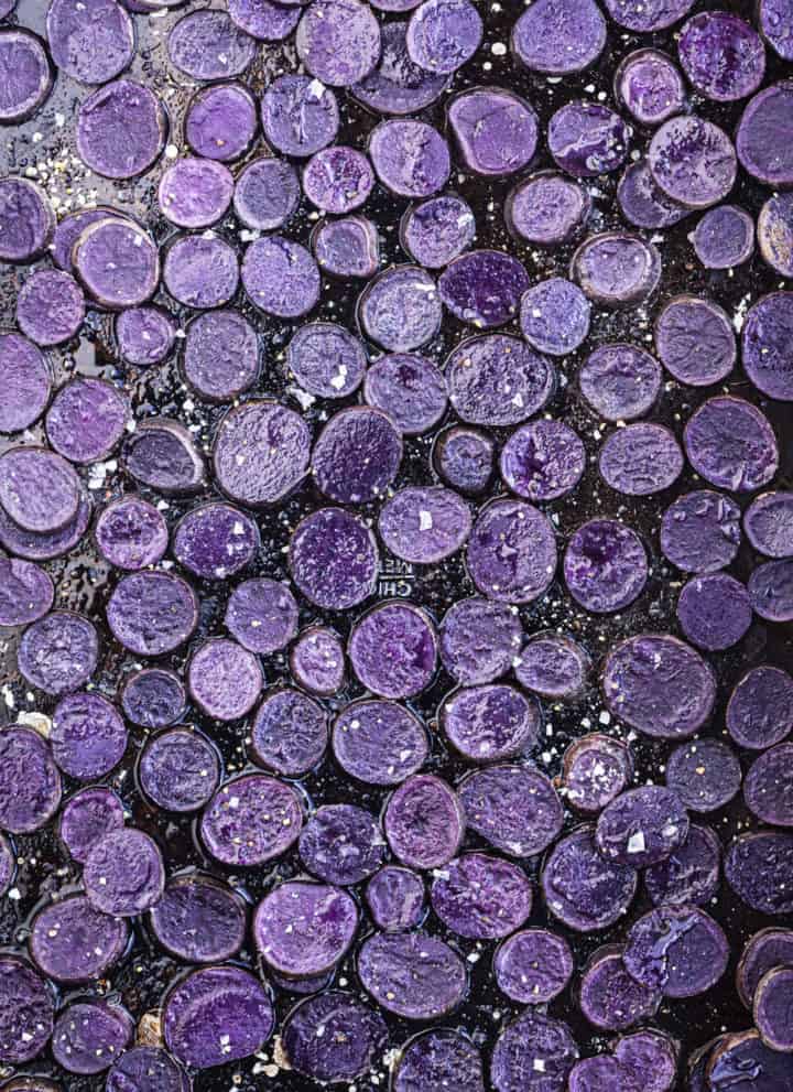 Small purple potatoes cut intro rounds, rounds are on a black baking sheet sprinkled with salt and overhead shot.