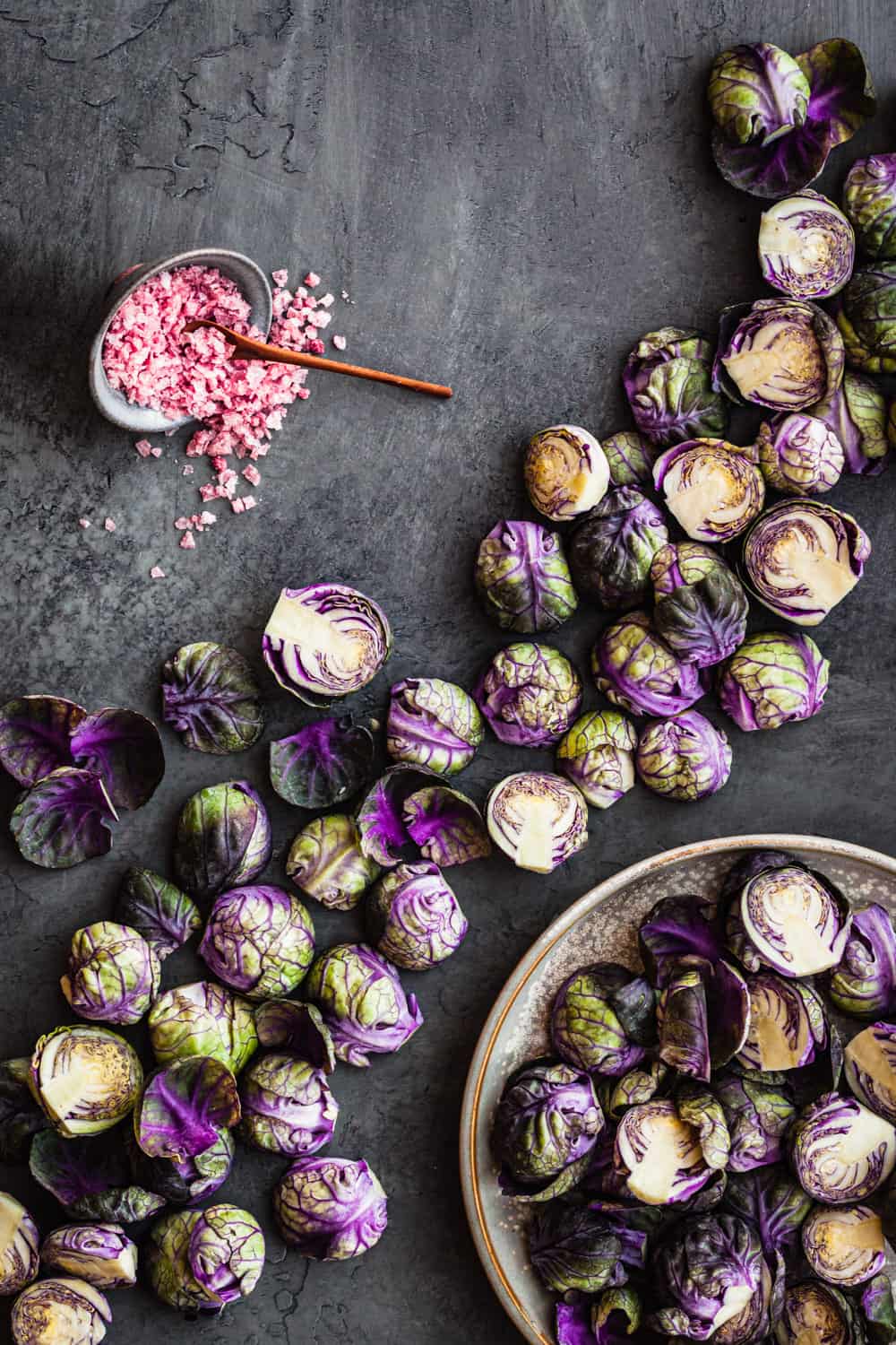 Purple Brussels sprouts! Some in a light bowl, some on a grey background, and a little bowl with purple salt; overhead shot.