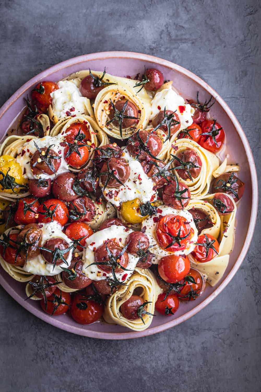 Different shades of red and yellow cherry tomato confit on top of pasta with burrata in a pink plate, overhead shot.