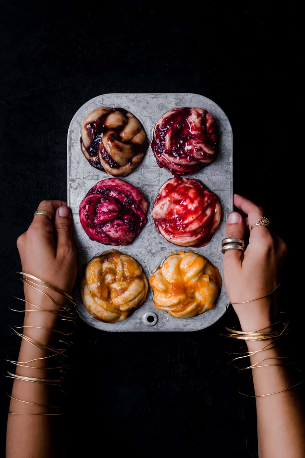 Daniela Gerson wearing jewelry and holding a silver muffin tin filled with challah babka buns, all with different color and type of jam; overhead shot.