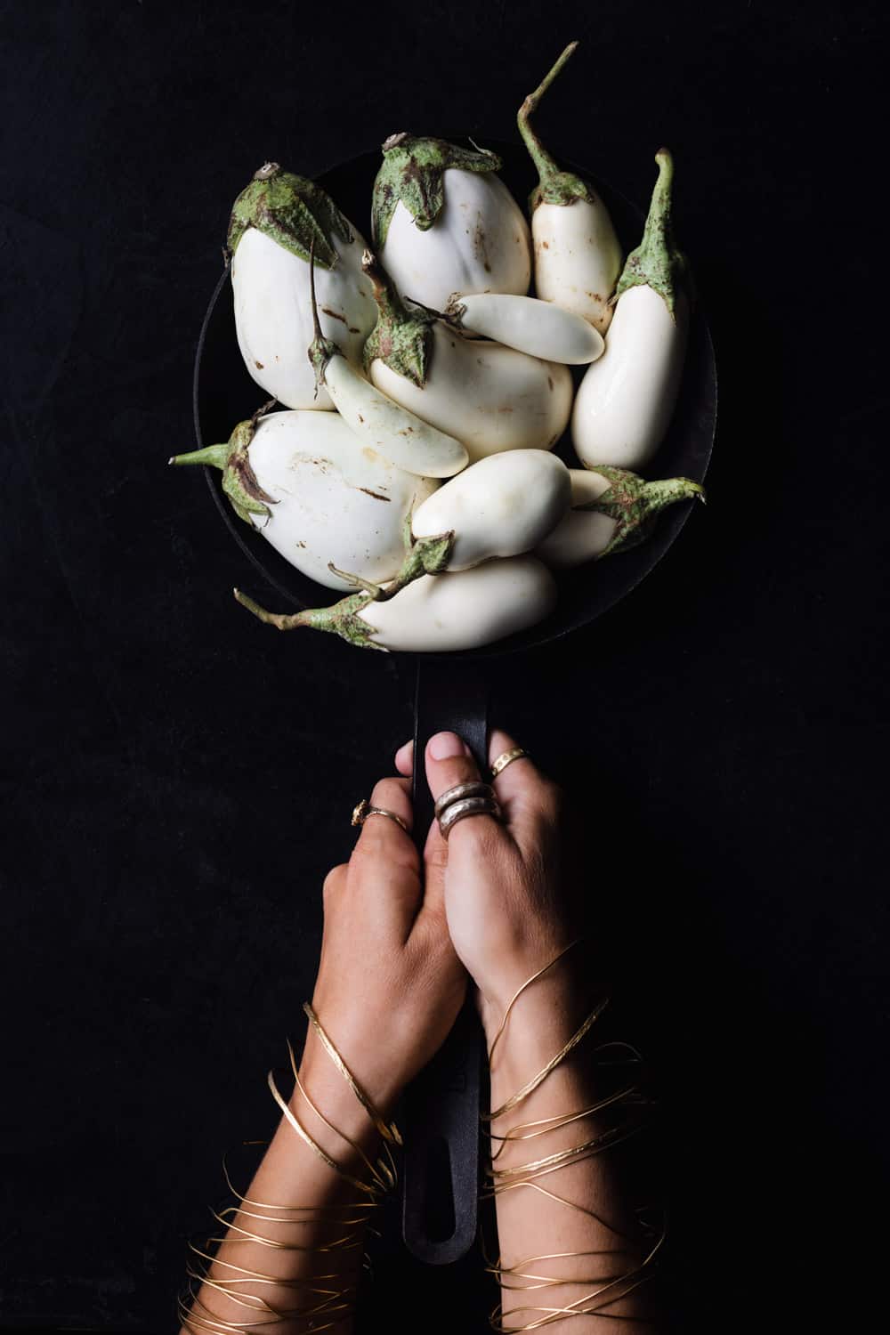 A cast iron skillet filled with different varieties of white eggplants, carried by Daniela Gerson wearing jewelry on a black background.