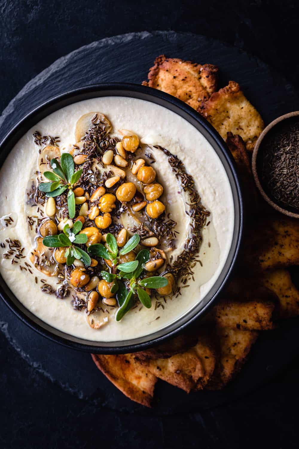 Hummus with tahini topped with cumin seed oil, garlic, crispy garbanzo beans, and fresh herbs; served with crispy pita chips; overhead shot.