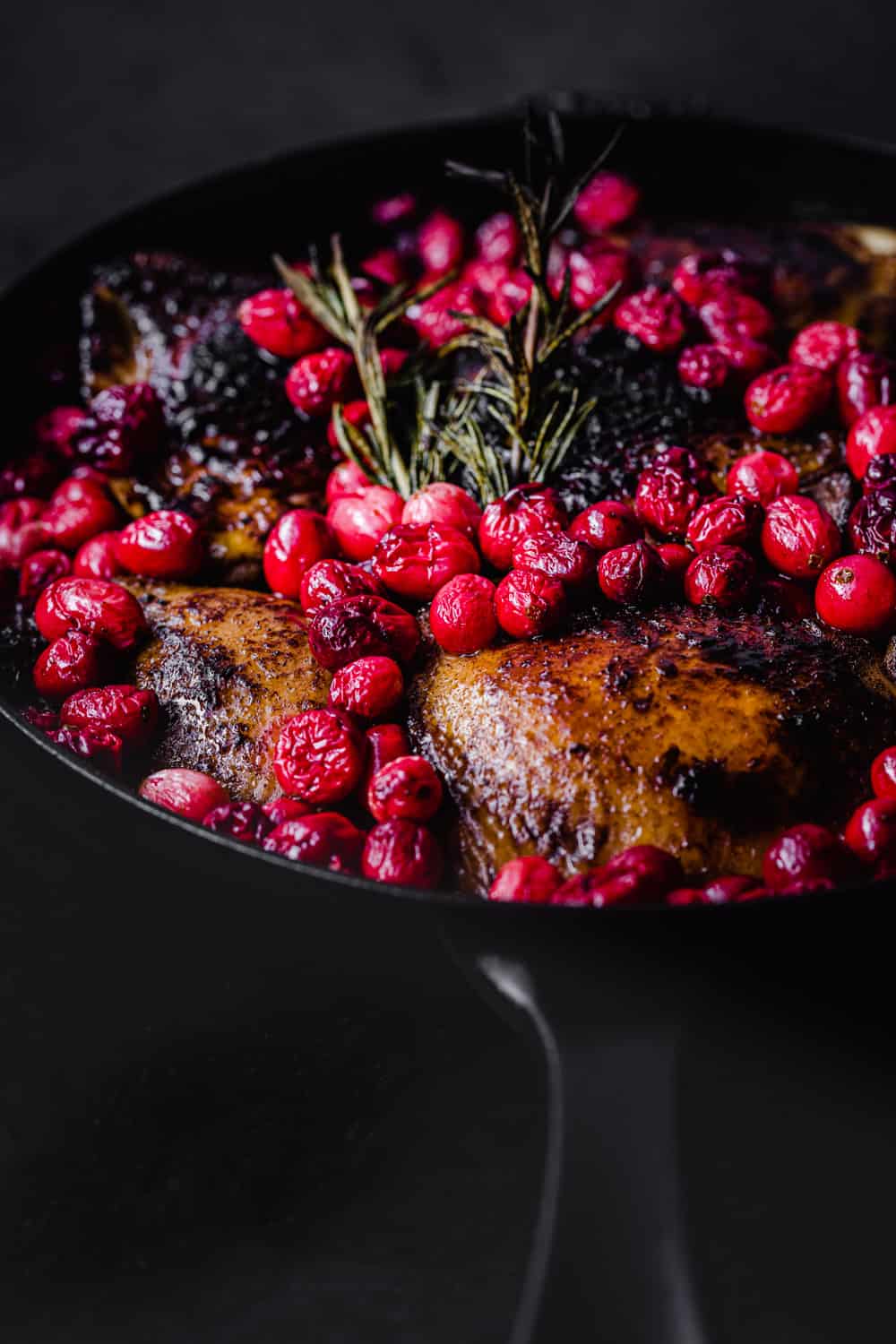 cranberry chicken with rosemary post oven in cast iron skillet; side angle shot on a black background.