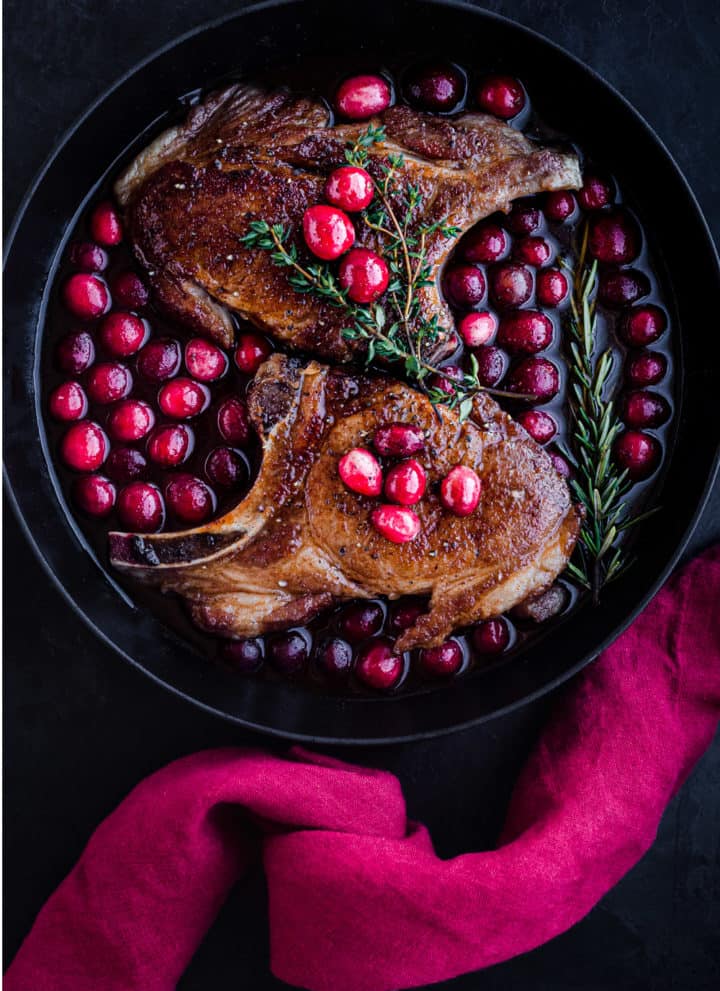 Post sear and pre-oven pork chop overhead shot; chops are with the cranberry sauce in a cast iron skillet with a red linen on the handle; black background.