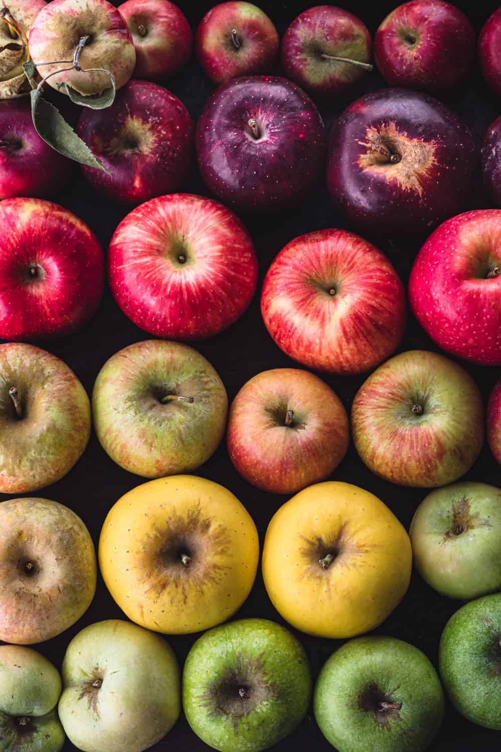 Apples arranged like a rainbow! There's red, purple, pink, yellow, green and hues in between; on a black background.