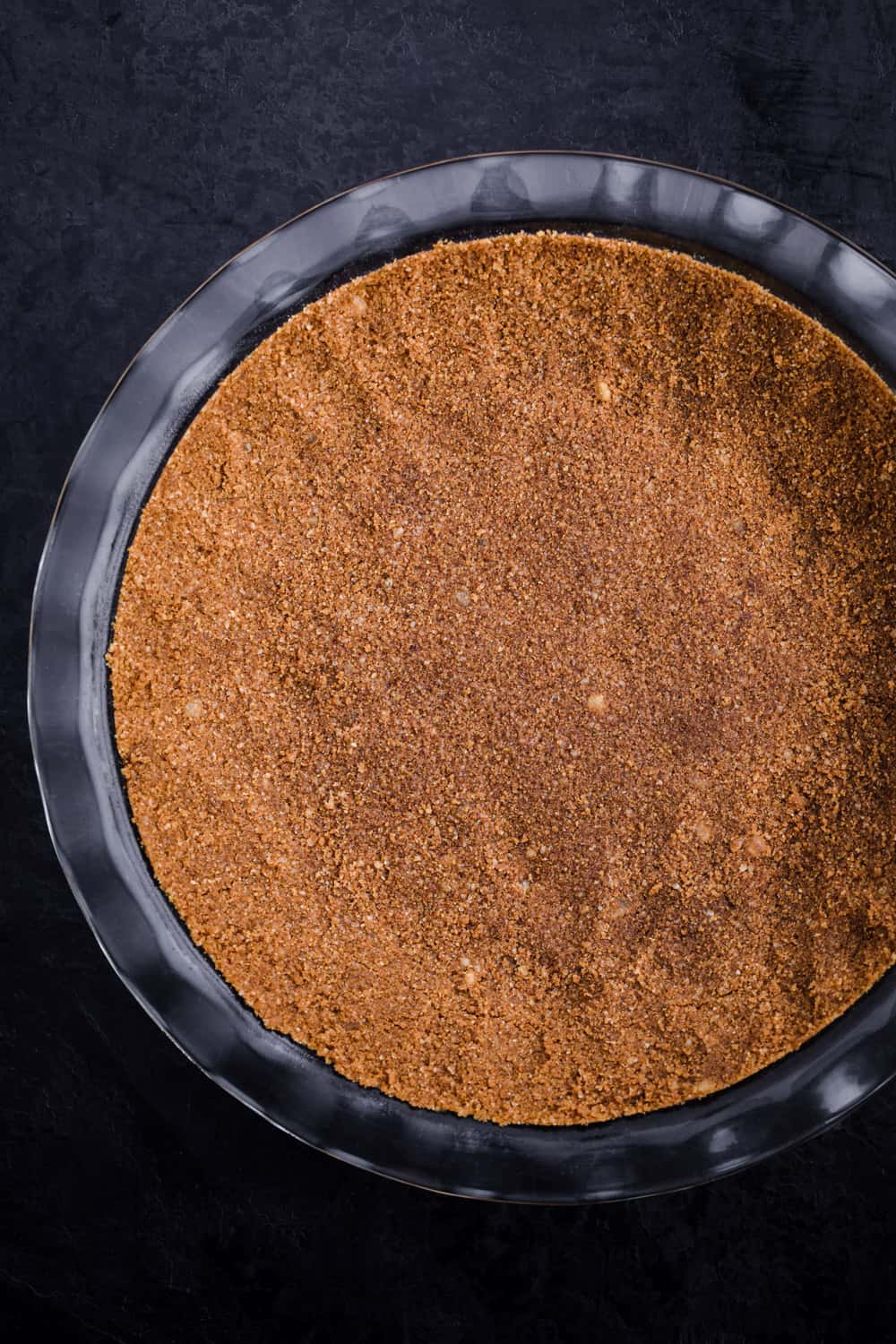 Gingersnap crust patted into a pie plate, all ready to be baked, overhead shot on a black background.
