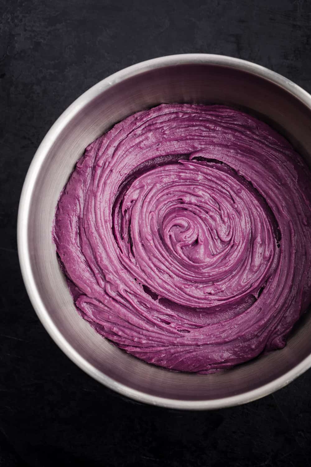 Purple sweet potato pie filling in a whisked together in a silver bowl; overhead shot on a black background.