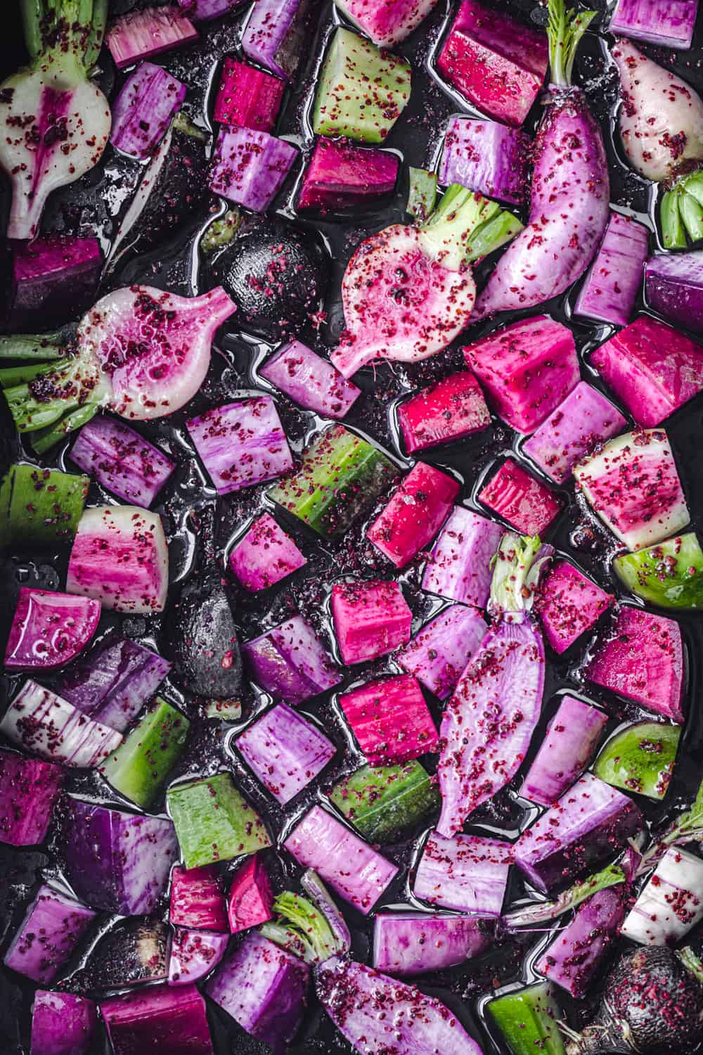 pink, purple, green and black raw radishes, cut into bite sized pieces and tossed with olive oil, sumac, salt and pepper; overhead shot.