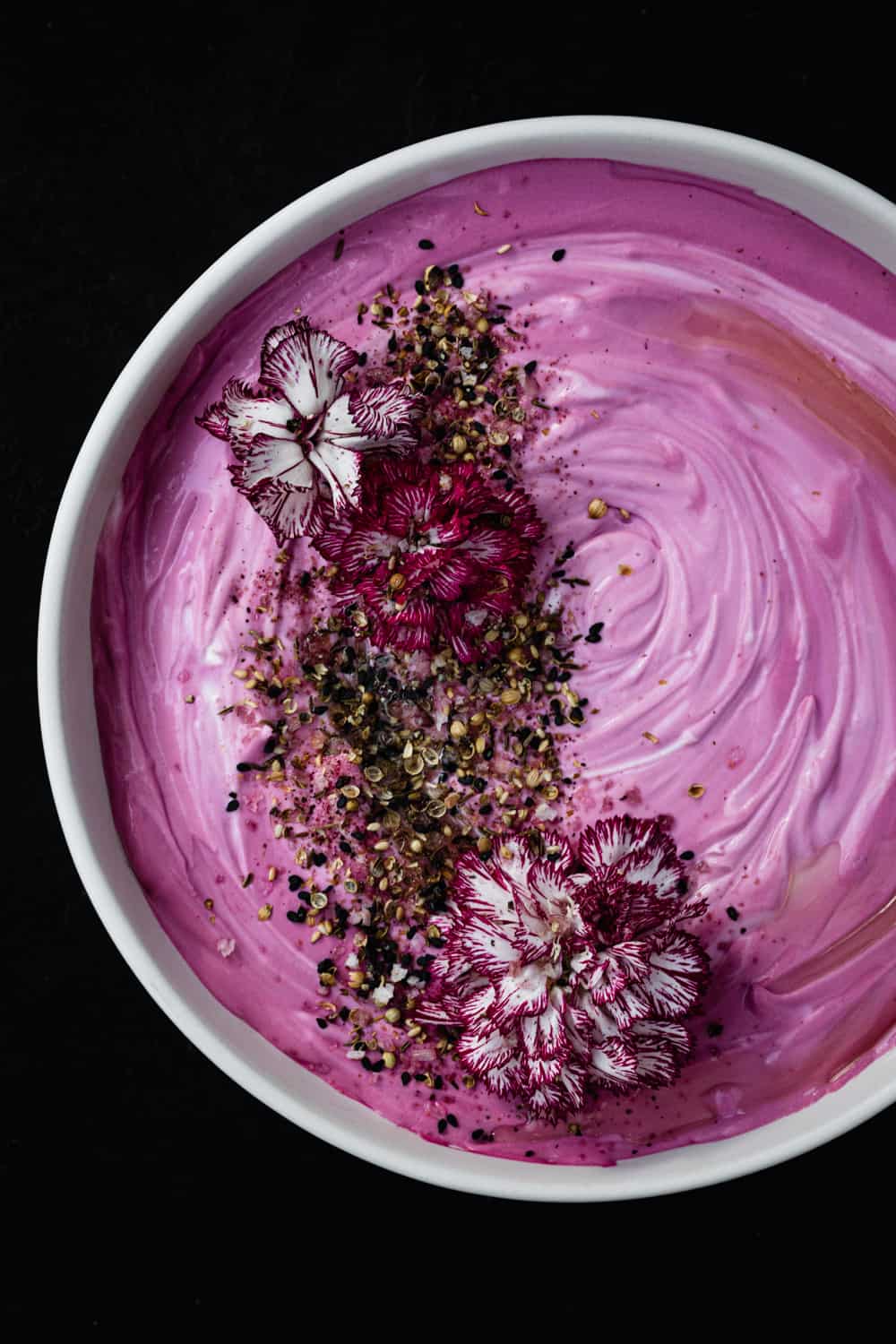 A vibrantly prink beet hummus topped with edible flowers, seeds and olive oil. Overhead shot on a black background.