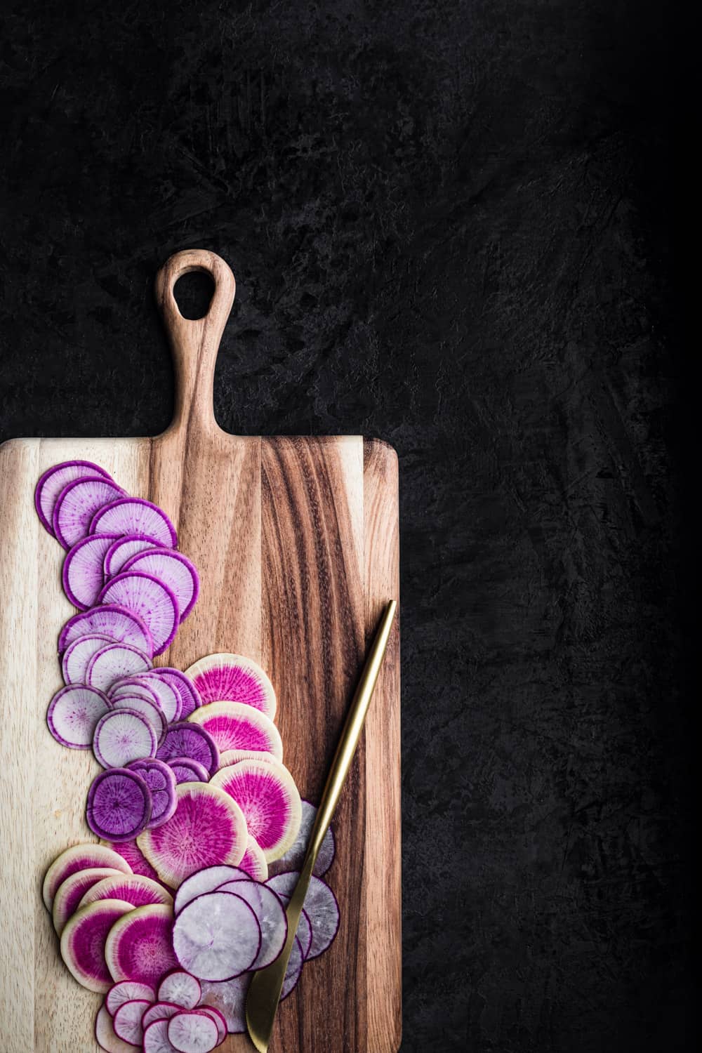 thinly sliced pink and purple radishes on a wooden cutting board with a gold knife; overhead shot on a black background.