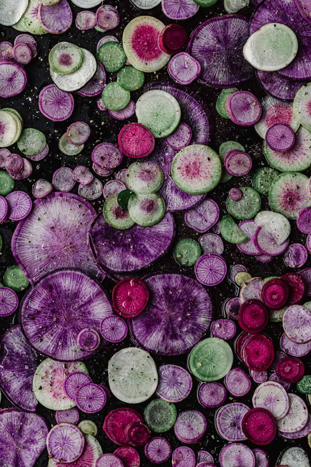 Pink, purple and green radishes thinly sliced into rounds on a baking sheet; overhead shot.