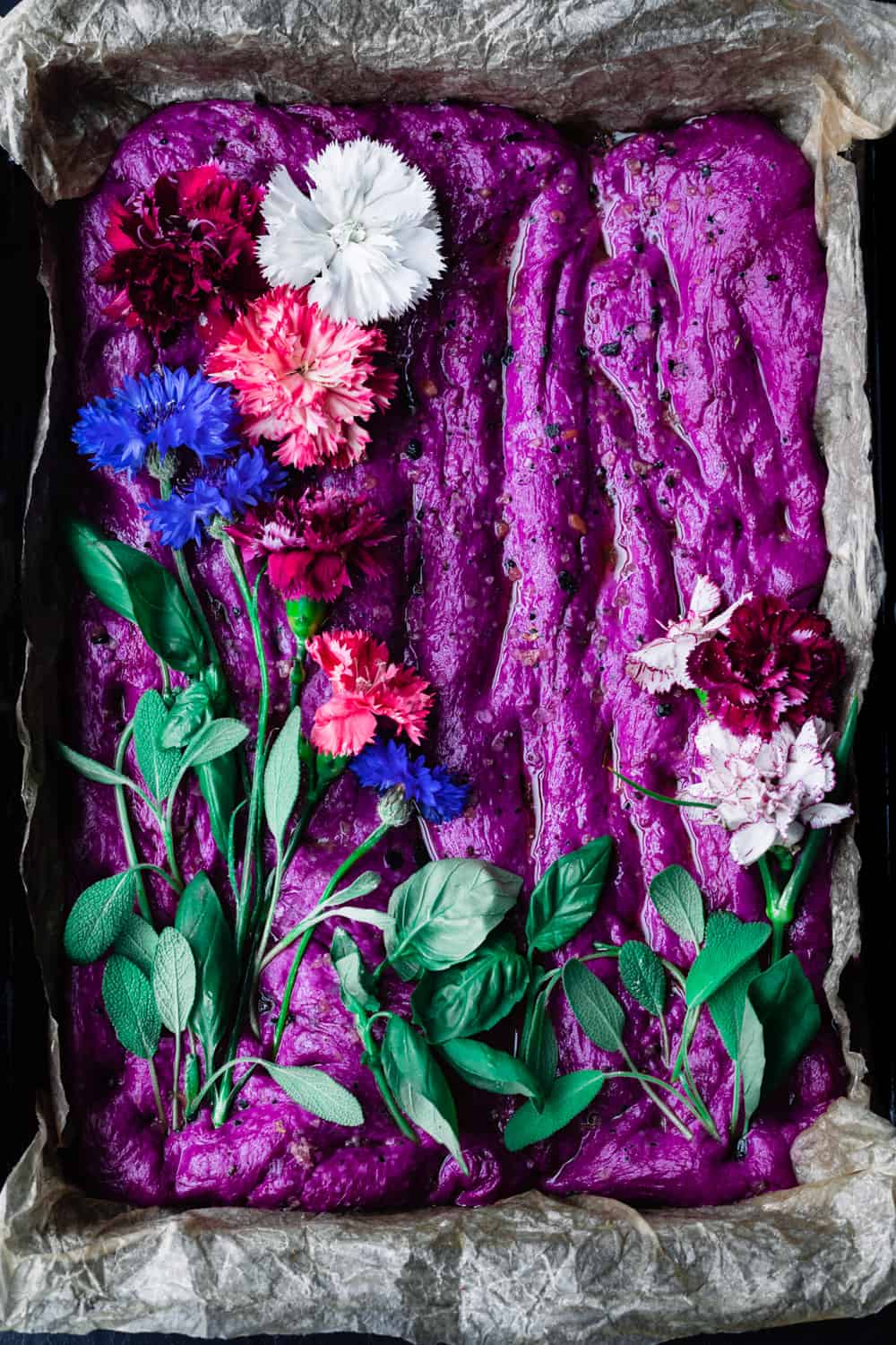raw focaccia dough in a baking pan topped with edible flowers and herbs; overhead shot.