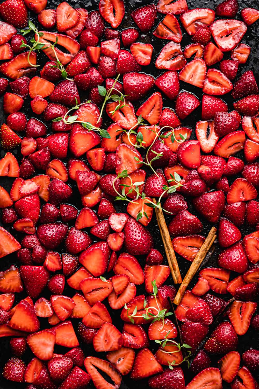 sliced strawberries tossed with sugar and topped with cinnamon, vanilla sticks and thyme on a baking sheet; overhead shot.