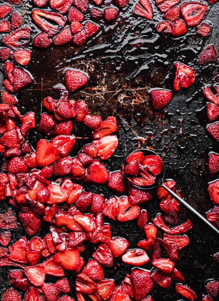roasted strawberries on a baking sheet with a black spoon; overhead shot.