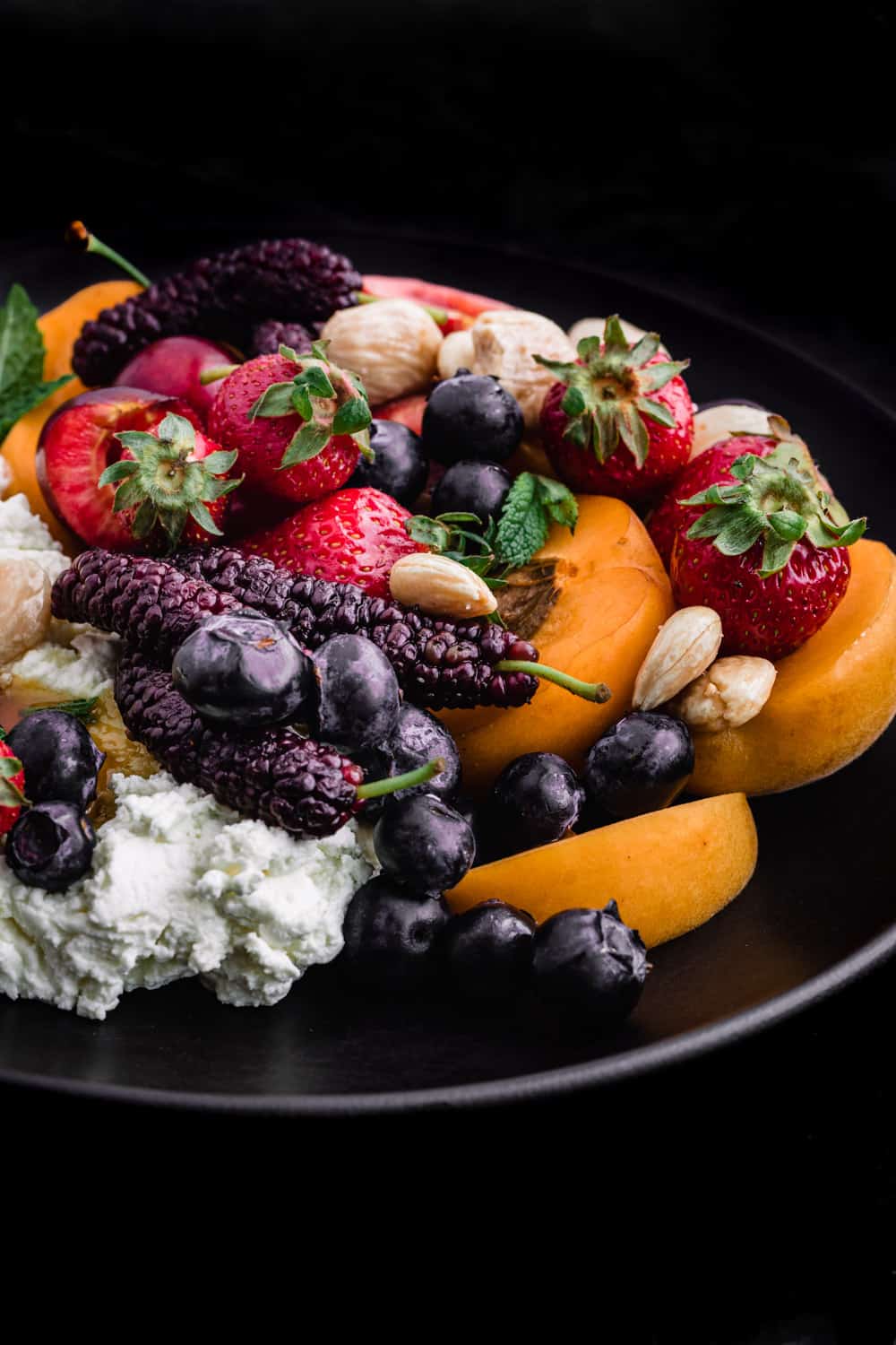 Single angle shot of Summer fruit with fresh cheese, almonds and honey on a black plate and background.