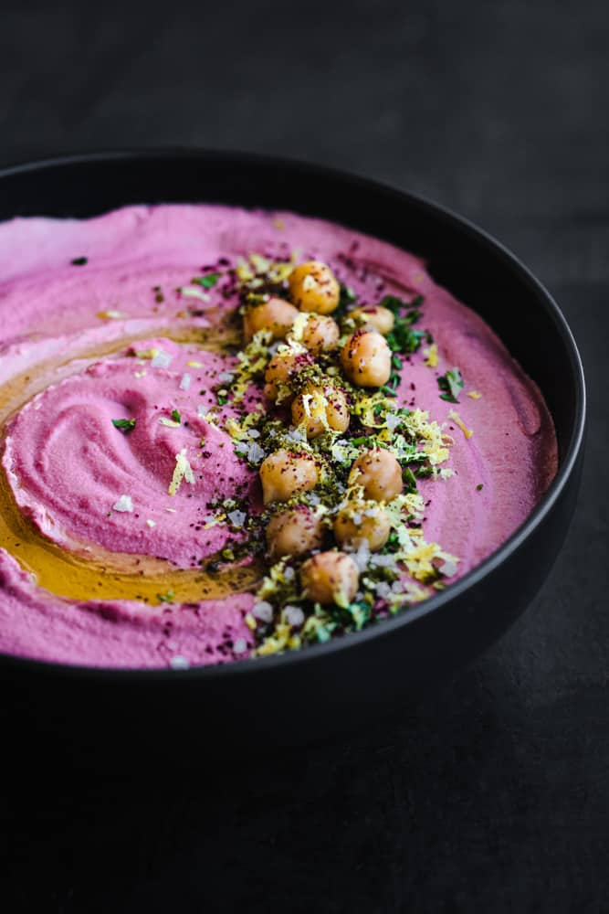 side angle shot of purple carrot hummus in a black bowl topped with garbanzo beans, herbs, spices and olive oil